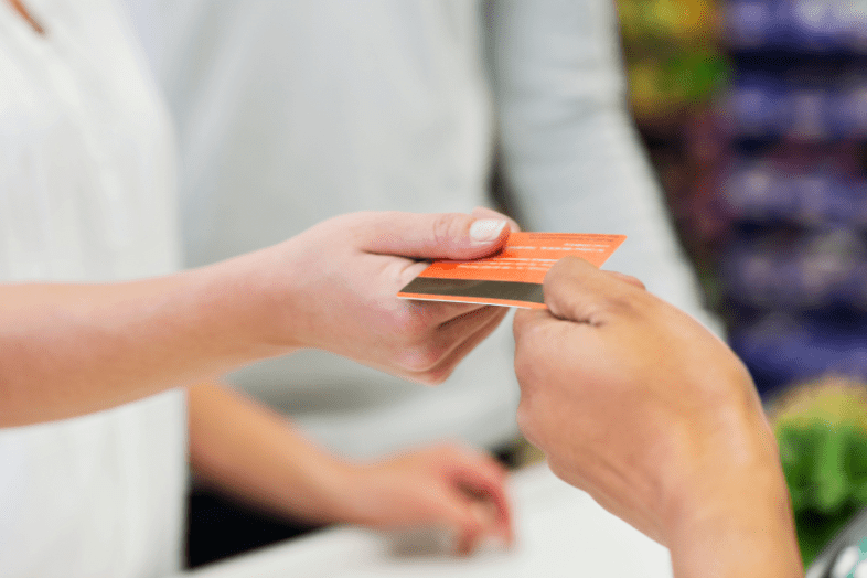 A closeup of a person’s hand giving an orange credit card to a cashier