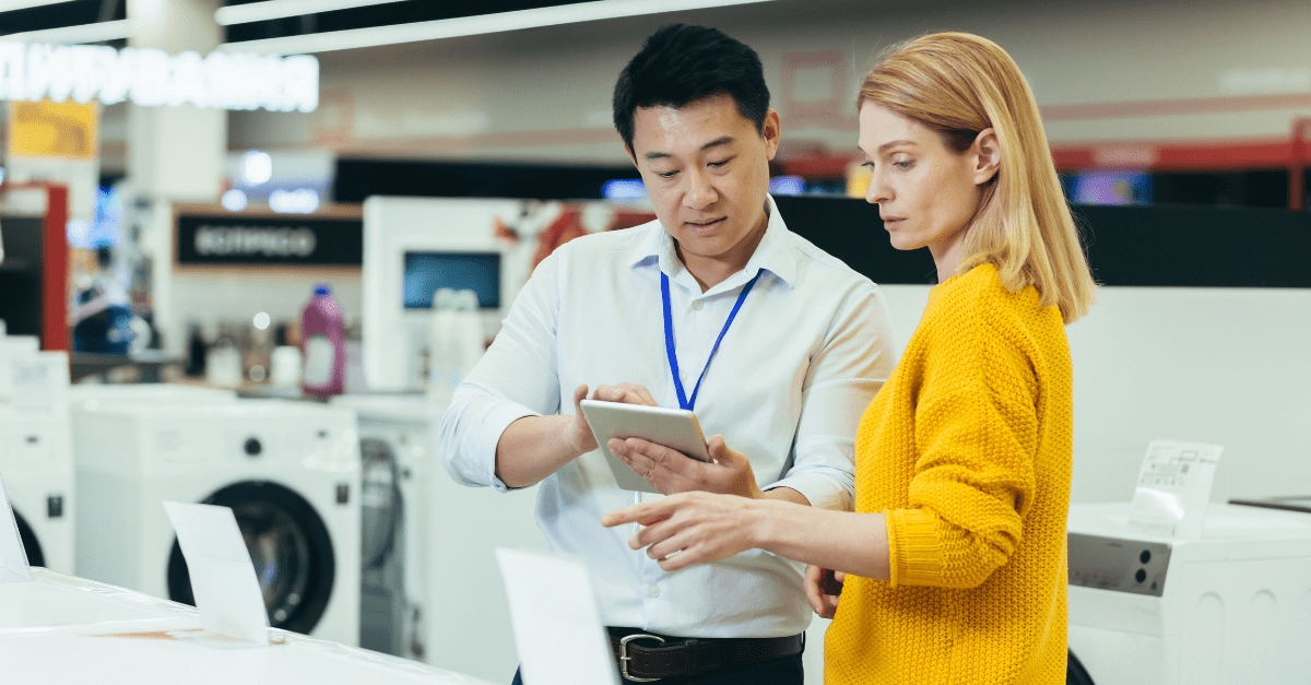 In an appliance showroom, a man and woman stand near washing machines. The man shows the woman information on a tablet while the woman points at a specific appliance.